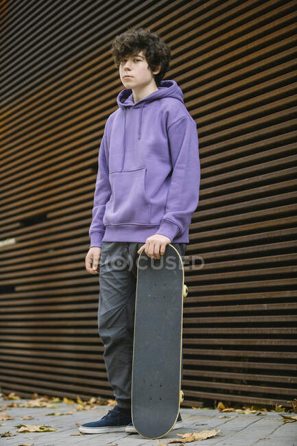 Corpo pieno di grave giovane maschio in abiti casual con skateboard guardando la fotocamera mentre in piedi vicino al muro sulla strada — Foto stock