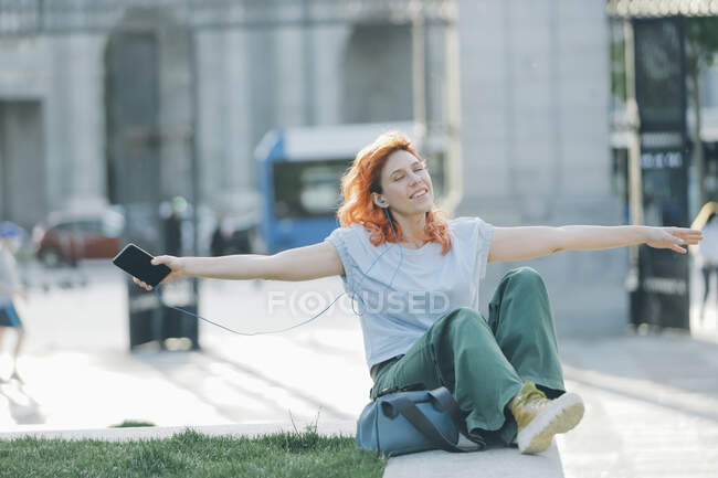 Mulher sonhadora com cabelos vermelhos sentada na rua e ouvindo música em fones de ouvido enquanto desfruta de músicas com olhos fechados e braços estendidos — Fotografia de Stock