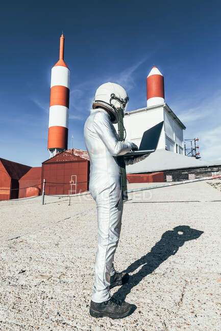 Astronauta macho de visión lateral en traje espacial navegando datos en netbook mientras está de pie fuera de la estación con antenas en forma de cohete - foto de stock