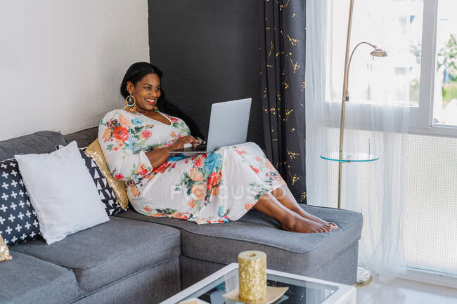 Femme afro-américaine adulte gaie dans une robe colorée lumineuse élégante et des boucles d'oreilles assises sur un canapé et un ordinateur portable de navigation tout en passant du temps libre à la maison — Photo de stock