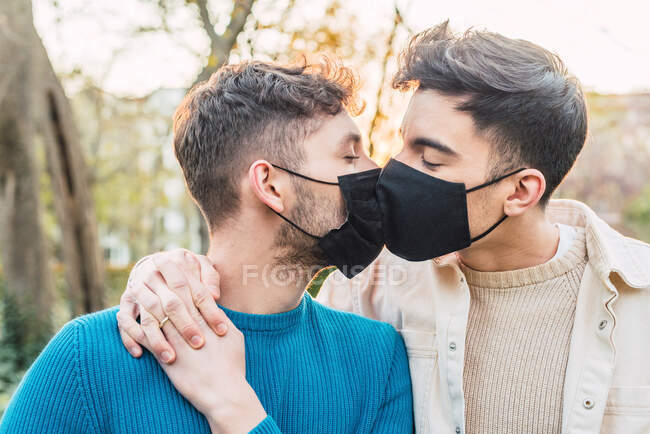 Amar a la pareja LGBT de hombres con máscaras protectoras abrazándose en el parque durante la epidemia de coronavirus y besándose - foto de stock