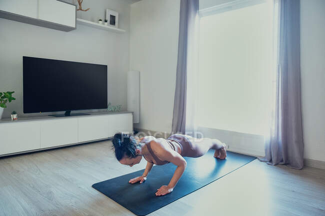 Femme en tenue de sport debout dans la pose de Phalakasana tout en pratiquant le yoga sur tapis dans la chambre de la maison le jour ensoleillé — Photo de stock