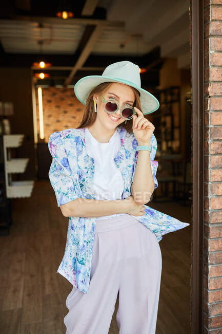 Веселая молодая женщина в модной одежде в солнечных очках смотрит в камеру на грубую стену в городе — стоковое фото