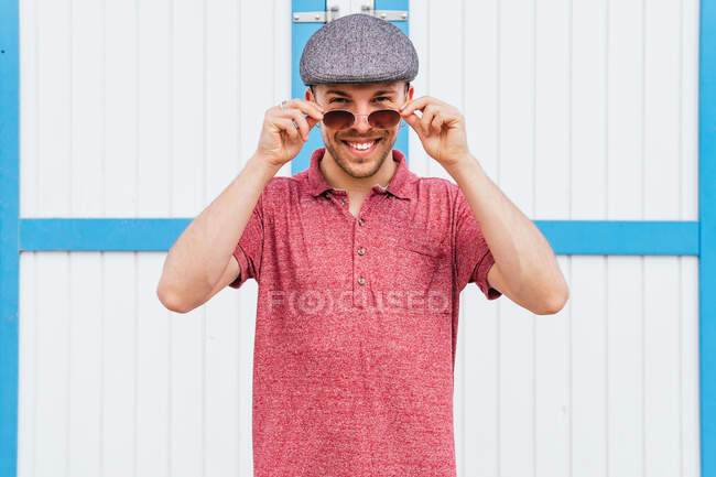 Молодой бородатый парень в обычной красной рубашке для поло и кепке смотрит на камеру в солнечных очках и улыбается дружелюбно, стоя у стены на улице в летний день — стоковое фото