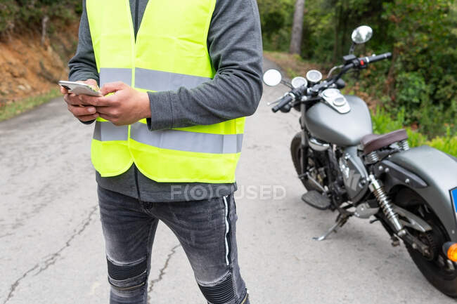 Crop motociclista maschio in gilet messaggistica sul telefono cellulare mentre in piedi vicino moto rotto vicino boschi verdi lussureggianti — Foto stock
