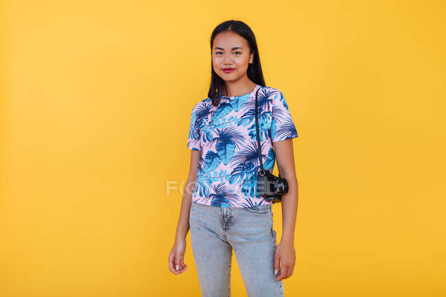 Mujer asiática feliz en camiseta con estampado de hoja tropical con cámara fotográfica sobre fondo amarillo en estudio - foto de stock