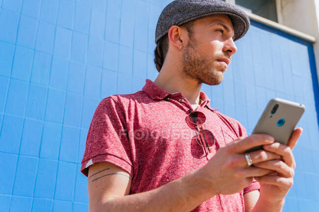 From below thoughtful young bearded hipster guy in casual polo shirt and cap browsing mobile phone while standing against blue wall in sunlight looking away — Stock Photo