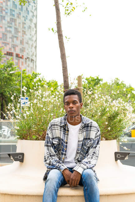Неэмоциональный афро-американский стильный мужчина сидит на скамейке с розетками для зарядки устройств в городе и смотрит в сторону — стоковое фото