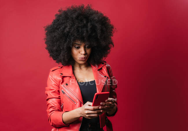 Femme afro-américaine excitée avec coiffure afro et lèvres boudantes naviguant sur téléphone mobile sur fond rouge en studio — Photo de stock