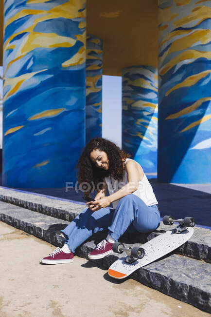 Full body side view of female in casual clothes sitting on stairs near skateboard browsing on smartphone on sunny street during training — Stock Photo