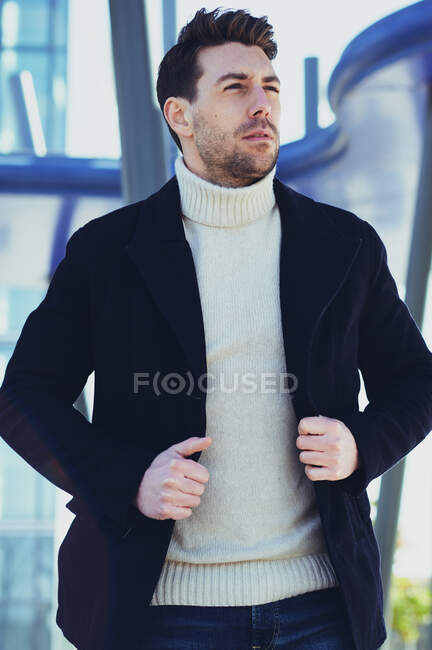 Взрослый бородатый мужчина в свитере и пальто прогуливается, глядя в город днем — стоковое фото