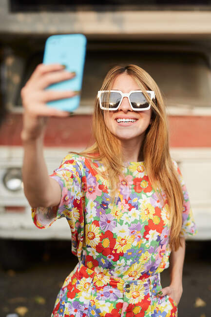Mujer alegre en ropa de verano de moda y gafas de sol de pie en la calle y tomando uno mismo tiro en el teléfono móvil - foto de stock