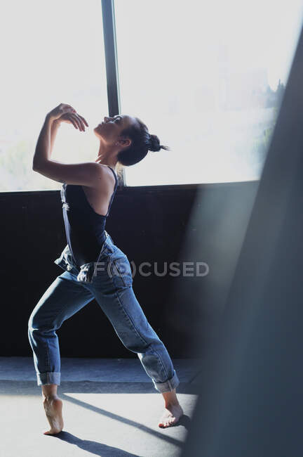 Young barefoot female in jeans with hair bun dancing while looking up on floor with shadows in sunlight — Stock Photo