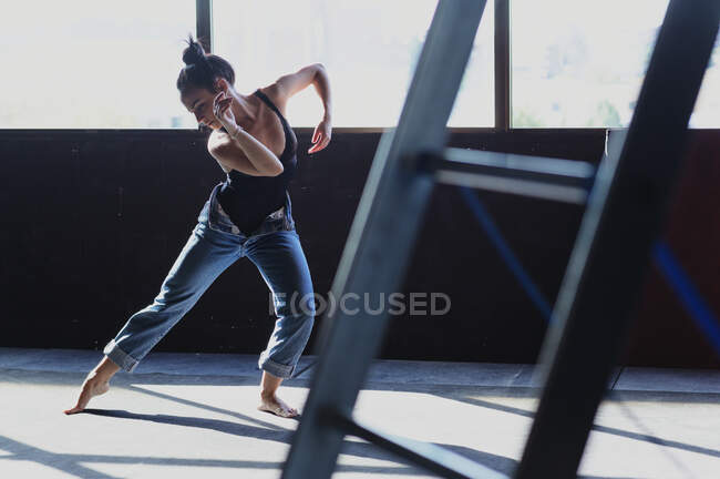 Молода босоніжка в джинсах з зачіскою бандою танцює, дивлячись вниз на підлогу з тінями на сонячному світлі — стокове фото