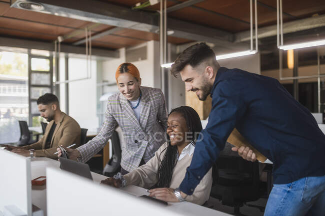 Grupo de diversos compañeros de trabajo alegres que se reúnen en la mesa con el ordenador portátil discutiendo proyecto mientras trabajan juntos en el espacio de trabajo contemporáneo - foto de stock