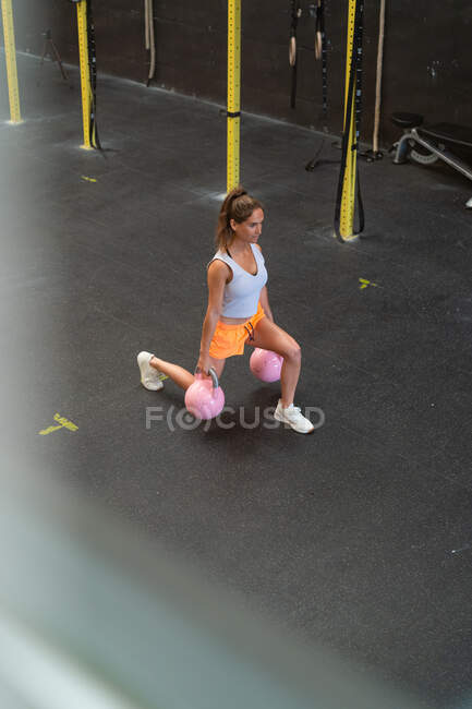 Высокий угол полного тела сильной спортсменки, делающей упражнения с тяжелыми гирями во время тренировки в тренажерном зале — стоковое фото