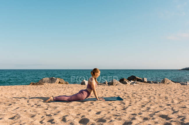 Vista lateral de la mujer haciendo yoga en Bhujangasana y estirando el cuerpo en la playa de arena en un día soleado - foto de stock