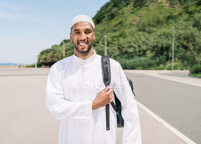 Happy Islamic man in traditional clothes adjusting backpack and looking away with smile while spending sunny summer day on beach — Stock Photo
