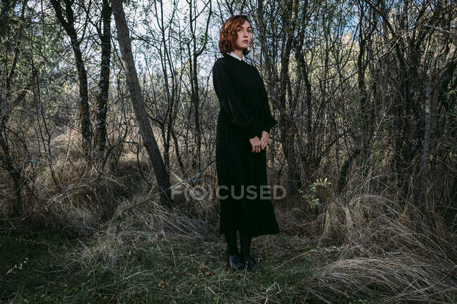 Mulher sem emoção vestindo vestido preto longo em pé na floresta de outono durante a celebração de Halloween — Fotografia de Stock