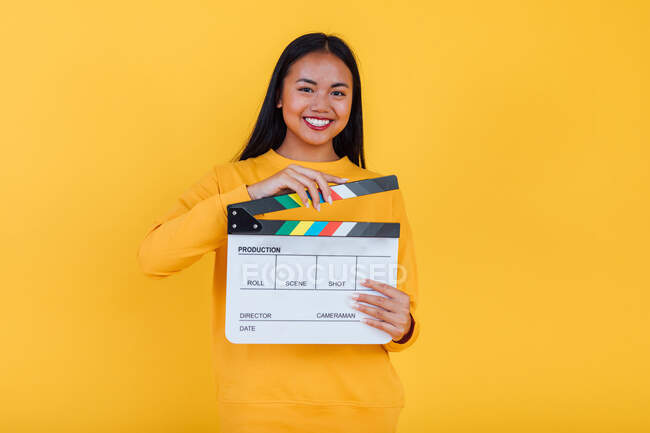 Fröhliche ethnische Frau steht mit Filmklöppel auf gelbem Hintergrund im Studio und blickt in die Kamera — Stockfoto