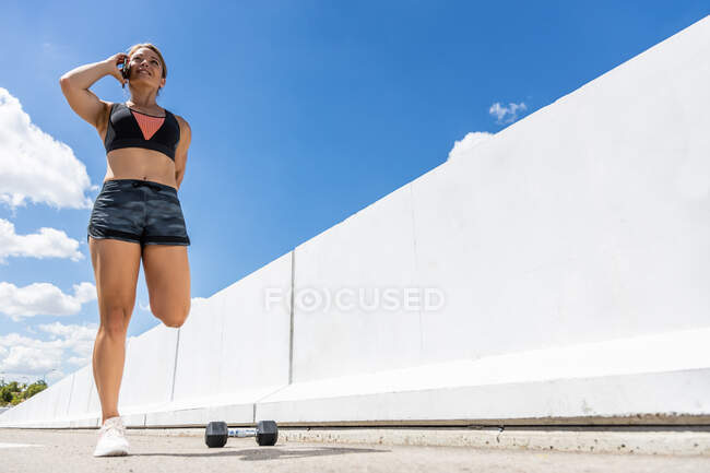 Junge Frau spricht nach einem Outdoor-Training mit ihrem Smartphone, während sie Muskeln dehnt, Seitenansicht — Stockfoto