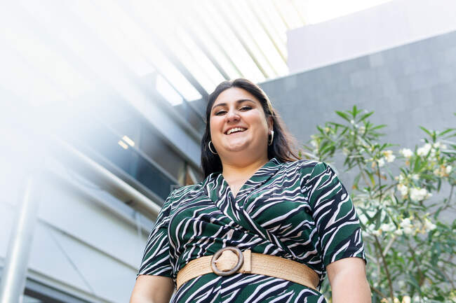From below of young stylish plus size female in elegant dress with geometric print and belt looking at camera and smiling friendly against modern urban background — Stock Photo