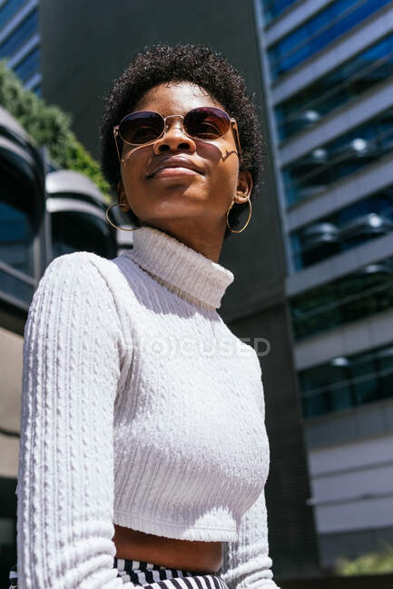 Нижче - позитивна молода афроамериканська жінка в модному одязі, яка дивиться у бік і насолоджується сонячним світлом, сидячи на лавці на сучасній вулиці міста. — стокове фото