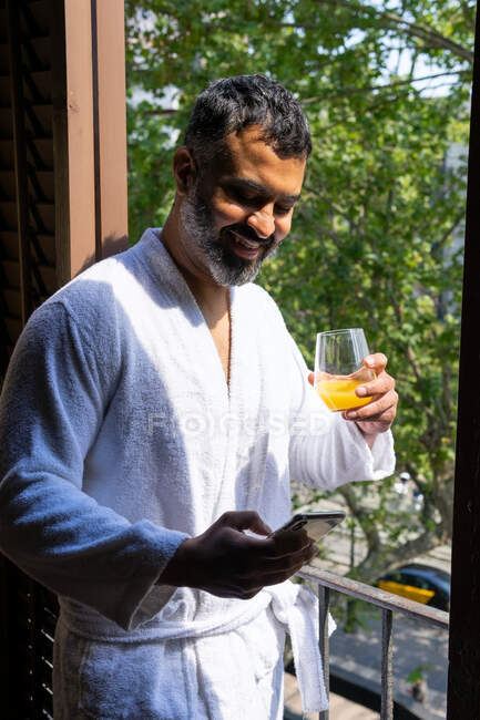 Улыбающийся взрослый мужчина в белом халате наслаждается свежим апельсиновым соком и просматривает смартфон, стоя на балконе отеля — стоковое фото
