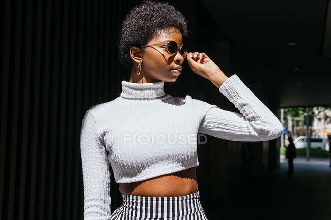 Jovem mulher negra em roupa elegante ajustando óculos de sol e olhando para longe no dia ensolarado na rua da cidade — Fotografia de Stock