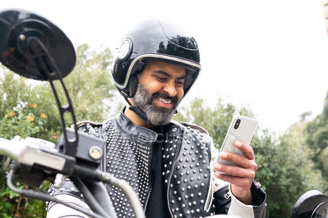 Бородатый велосипедист в модной кожаной куртке с заклепками и защитным шлемом просматривает телефон, сидя на мотоцикле, припаркованном рядом с пышным зеленым лесом — стоковое фото