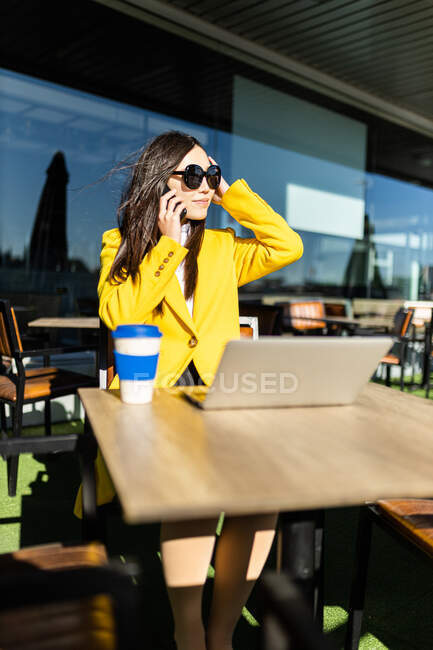 Souriant asiatique femme d'affaires avec manteau jaune assis à une table prenant un café avec son téléphone intelligent et ordinateur portable — Photo de stock