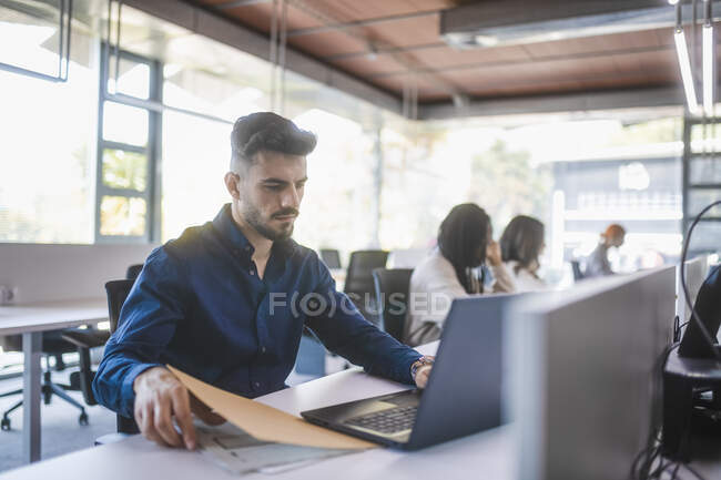 Empregado masculino concentrado sentado à mesa com laptop e lendo documentos enquanto trabalhava em um escritório espaçoso com colegas desfocados — Fotografia de Stock