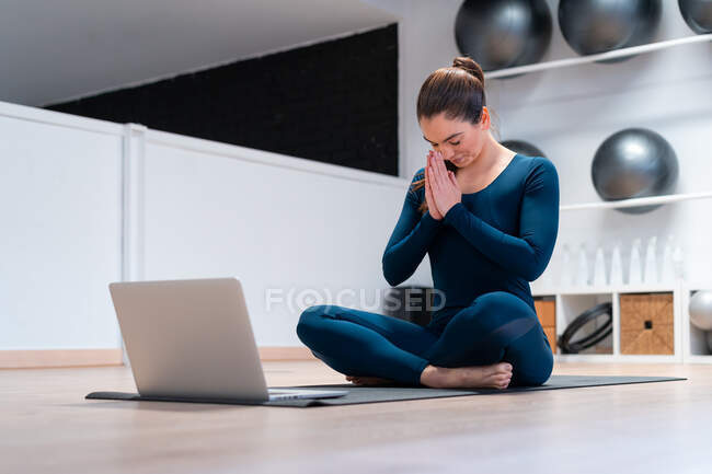 Full body of female instructor meditating with namaste hands and eyes closed while practicing yoga via video chat on laptop during online yoga class in fitness studio — Stock Photo