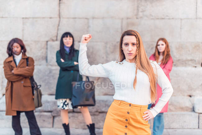 Mulher confiante mostrando bíceps enquanto está contra o grupo de mulheres multirraciais mostrando conceito de poder feminino — Fotografia de Stock