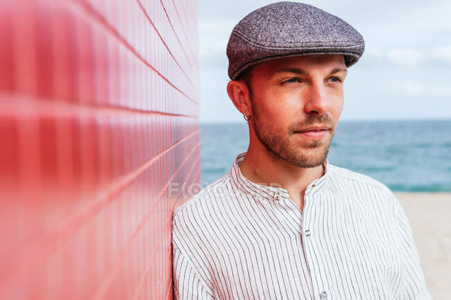 Thoughtful young bearded male in stylish striped shirt and hat standing looking away near red wall and enjoying sunny summer day on street — Stock Photo