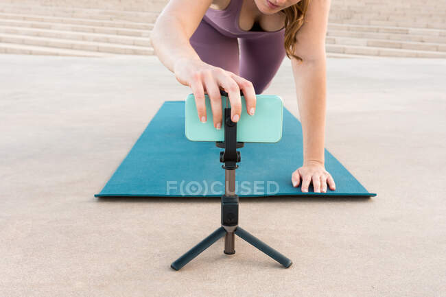 Ângulo alto da fêmea irreconhecível cortada que prepara o telefone móvel no tripé para fazer o ioga durante a lição em linha — Fotografia de Stock