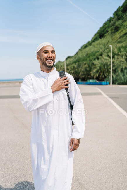 Hombre islámico feliz con ropa tradicional ajustando la mochila y mirando hacia otro lado con sonrisa mientras pasa el soleado día de verano en la playa - foto de stock