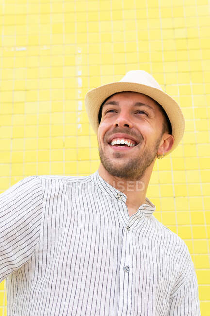 From below of happy young bearded hipster guy in trendy outfit and hat smiling while chilling alone against yellow wall in summer day — Stock Photo