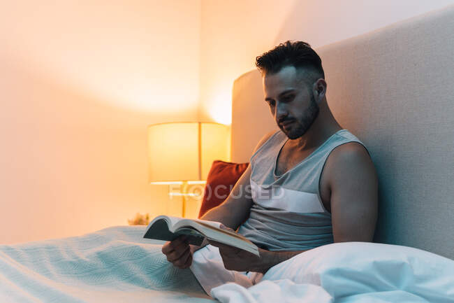 Задумчивый мужчина сидит на кровати под одеялом и читает интересную историю в книге перед сном — стоковое фото