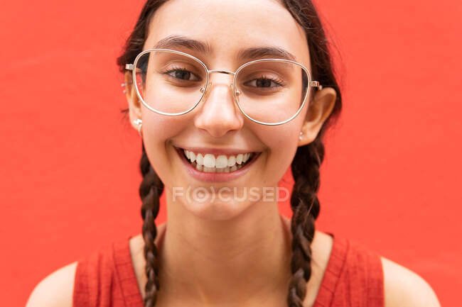 Alegre joven hembra en coletas peinado mientras mira a la cámara en el fondo rojo en la calle - foto de stock