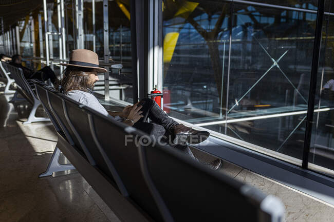 El tipo en el sombrero en el aeropuerto en la sala de espera sentado esperando su vuelo, con auriculares inalámbricos para escuchar música mientras charla con su teléfono inteligente, vista lateral - foto de stock