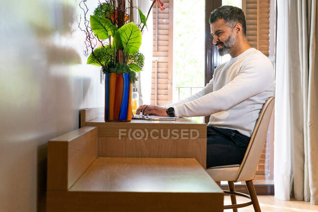 Vista lateral hombre de negocios étnico escribiendo en netbook sentado en la mesa en la habitación de hotel durante el viaje de negocios - foto de stock