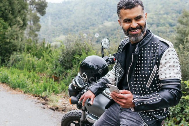 Side view of bearded male biker in trendy leather jacket with rivets and protective helmet browsing phone while sitting on motorcycle parked near lush green forest — Stock Photo