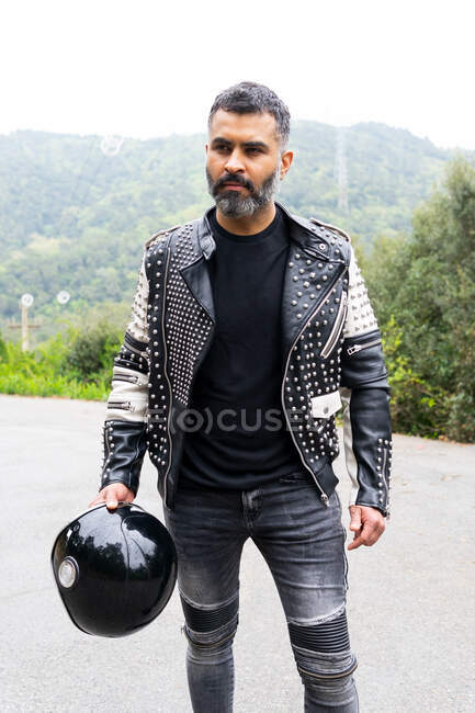 Serious brutal ethnic male biker in stylish leather jacket standing on asphalt road with helmet in hand near lush green trees growing in mountainous valley — Stock Photo