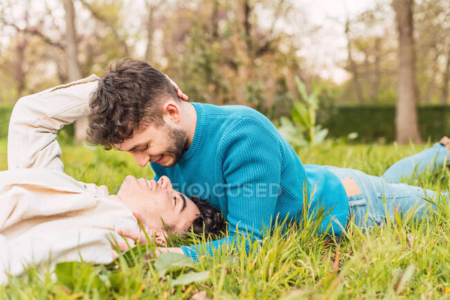 Vista lateral de la feliz pareja homosexual de hombres acostados en el prado en el parque y mirándose el uno al otro - foto de stock