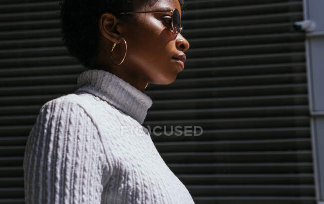Side view of young African American woman in stylish sweater and sunglasses looking away while standing in bright sunlight against black background — Stock Photo