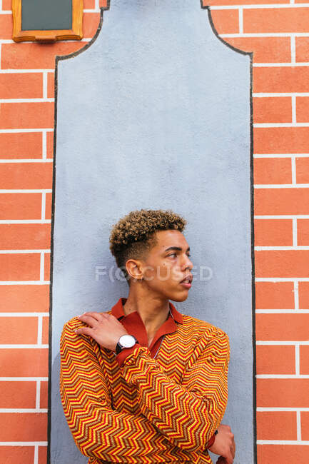 Jovem elegante pensativo étnico encaracolado cara na roupa da moda inclinando-se contra a parede de tijolo na rua urbana olhando para longe — Fotografia de Stock