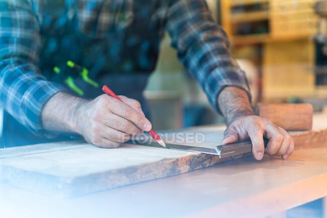 Crop adult male woodworker with pencil and ruler marking wooden board while working at workbench in carpentry workshop — Stock Photo