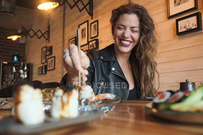 Улыбающаяся женщина ест вкусные суши в японском ресторане, сидя за деревянным столом — стоковое фото