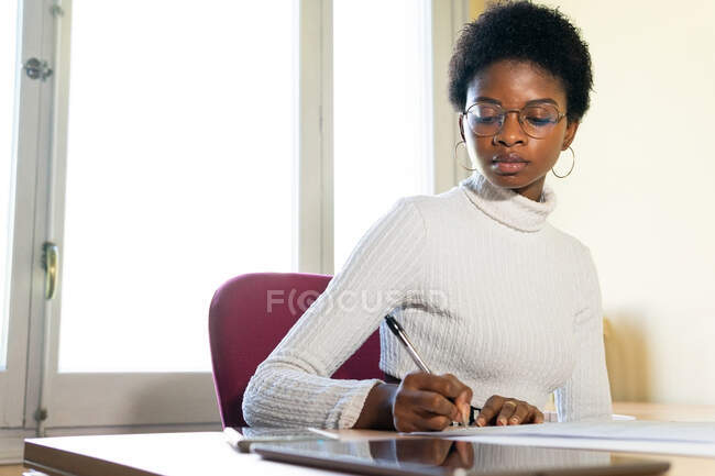 Médico negro escribiendo información en hoja de papel mientras prepara informe médico en la mesa en la oficina de la clínica moderna - foto de stock